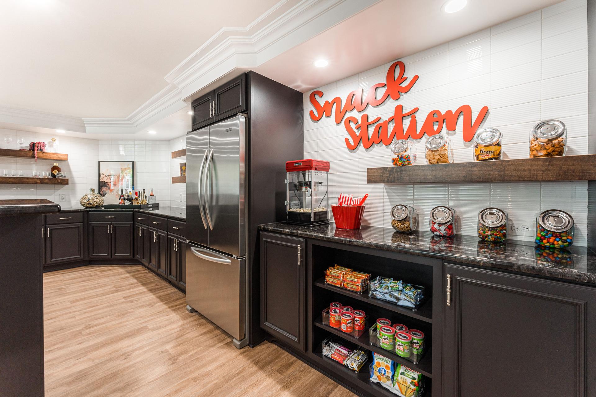Snack station wall — stainless French door refrigerator, open shelving with candy jars, popcorn machine, and custom neon sign — Geneva IL basement finish