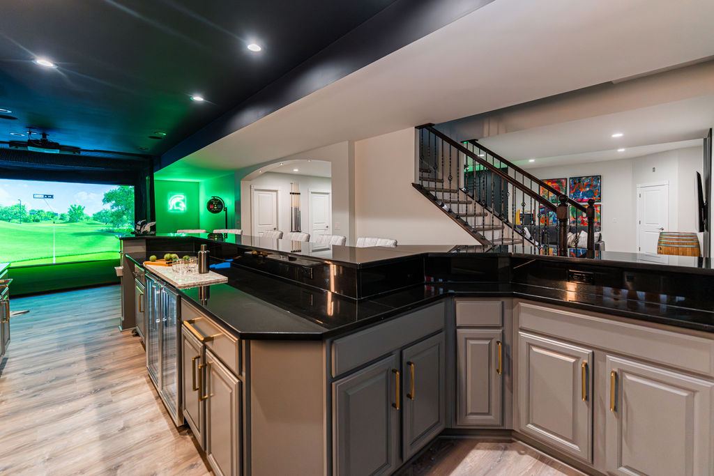 Hickory St. Charles — bar island overview with black granite, brass pulls, beverage refrigerator, and golf simulator beyond
